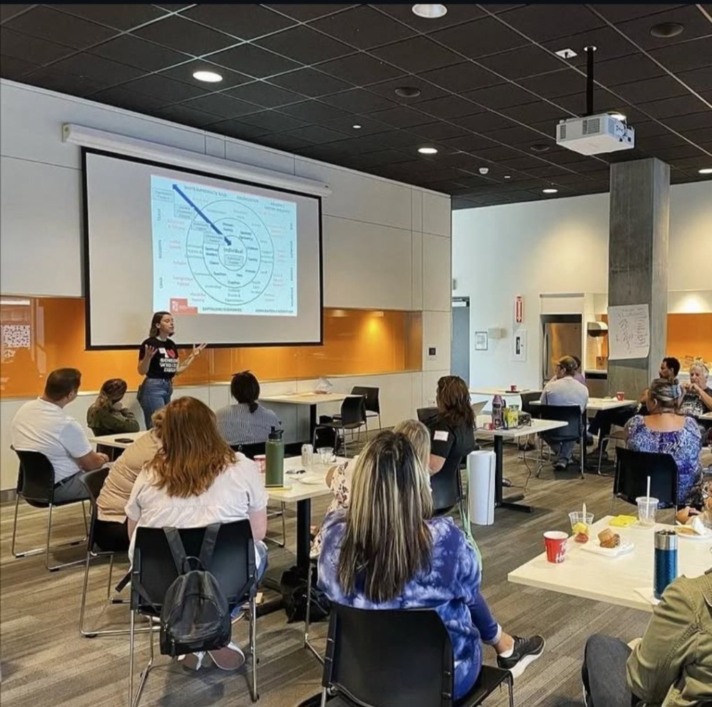 A facilitator (Jordyn) presenting to an audience in a workshop setting, with a projection screen displaying a diagram related to harm reduction.